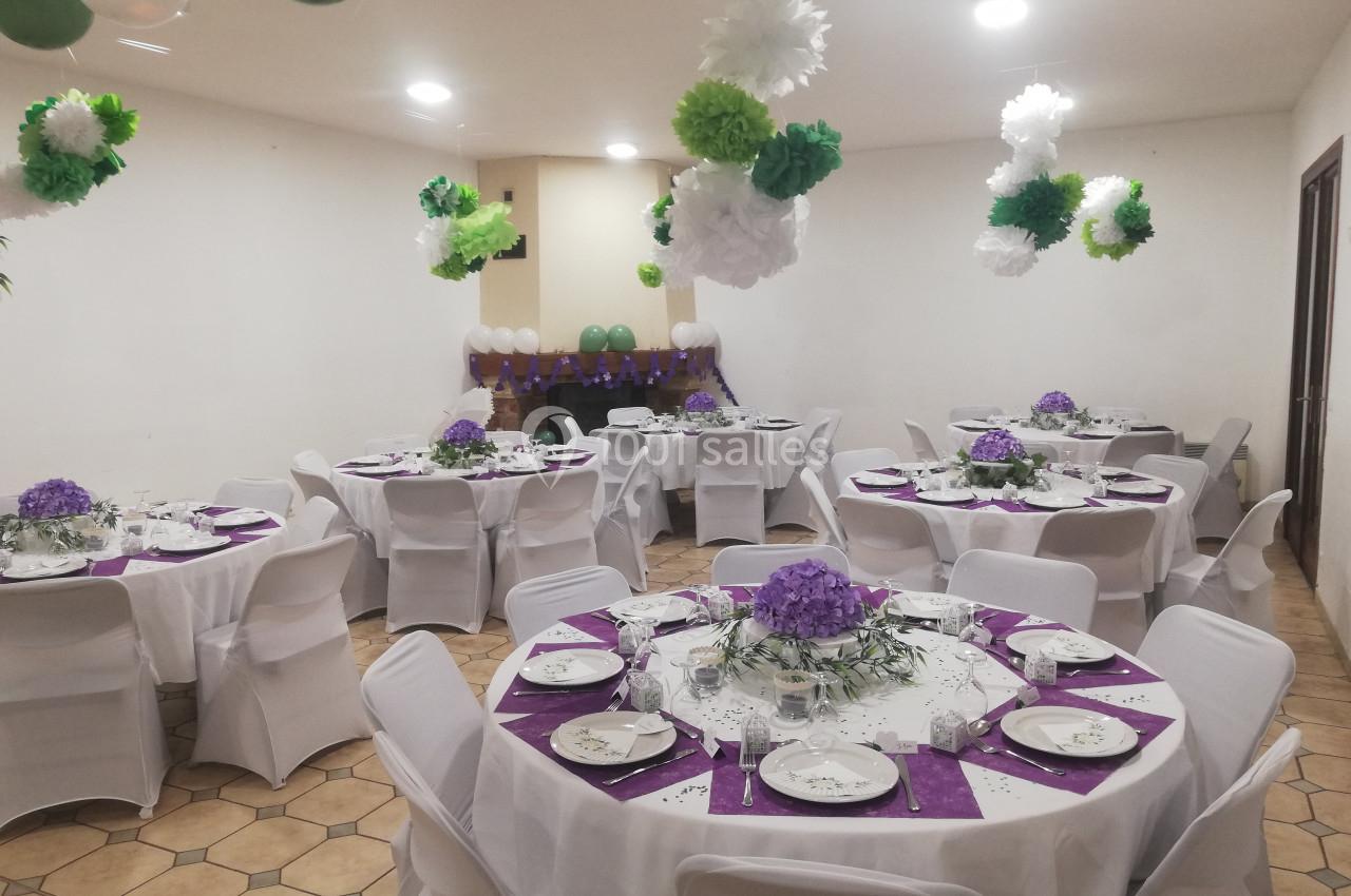 Salle décorée pour un événement avec des tables rondes dressées, nappes blanches, centres de table violets et guirlandes…