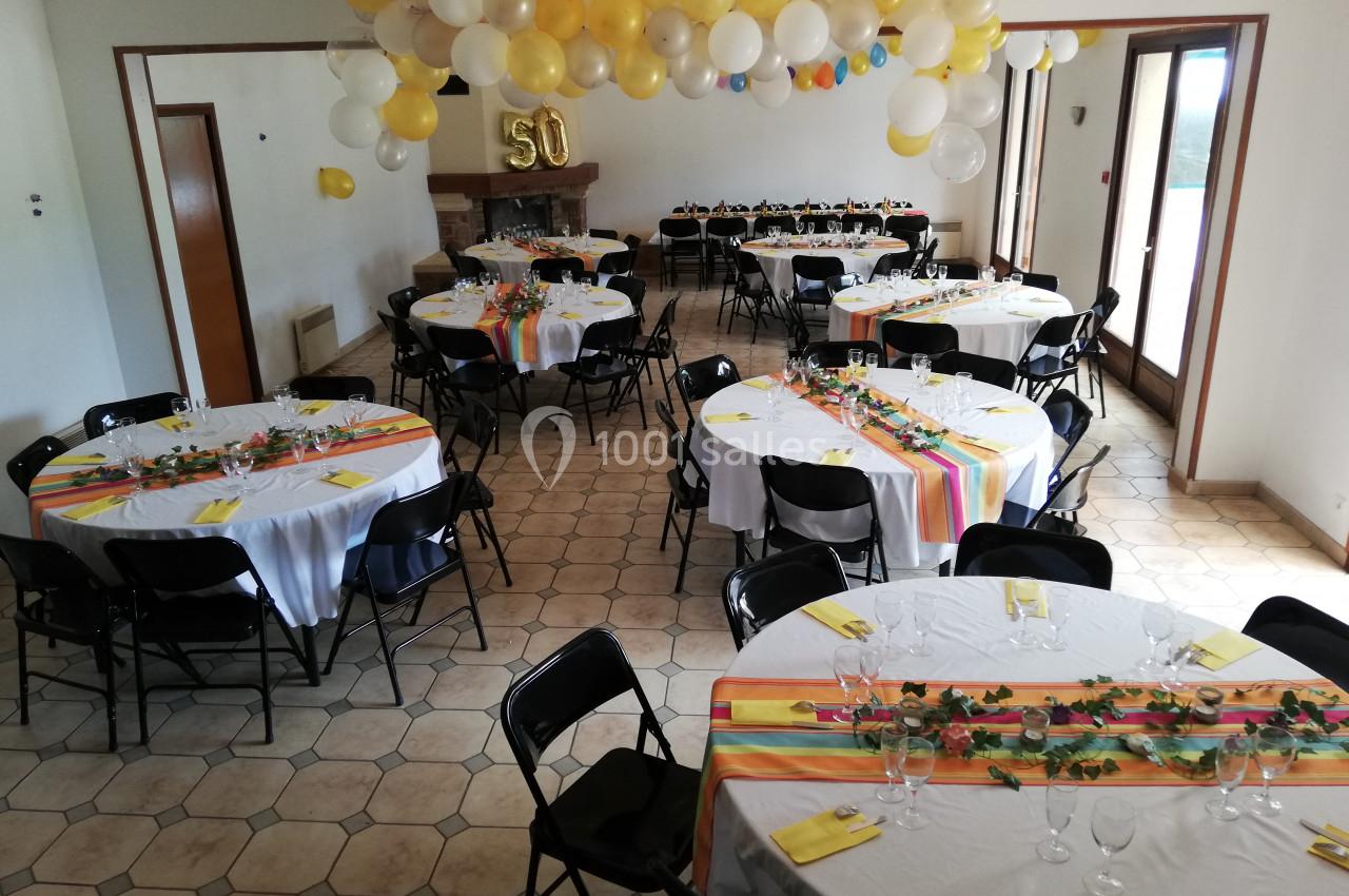 Salle décorée pour une fête avec des tables rondes dressées, des nappes blanches et des ballons suspendus au plafond.