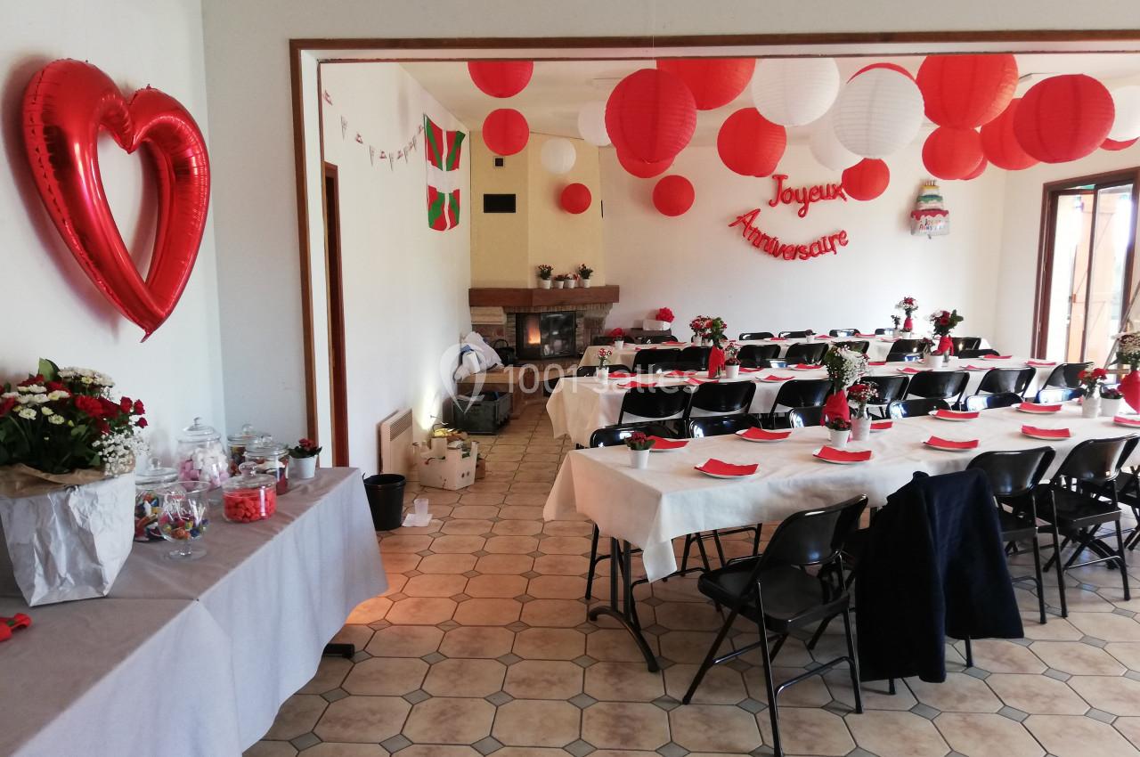Salle décorée pour un anniversaire avec tables dressées, ballons rouges et blancs, et une guirlande ’Joyeux Anniversaire’.