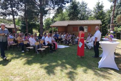 Des personnes assises en extérieur assistent à une cérémonie devant des tables décorées, près de chalets en bois.
