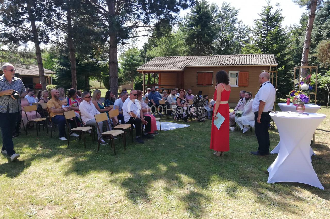 Des personnes assises en extérieur assistent à une cérémonie devant des tables décorées, près de chalets en bois.