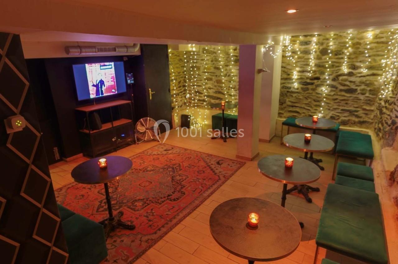 Salle cosy avec tables basses, banquettes, tapis rouge, murs en pierre et guirlandes lumineuses.