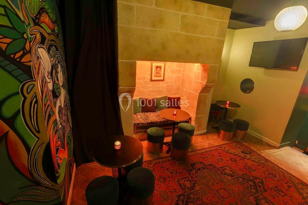 Espace cosy avec alcôve en pierre, tables basses, tabourets, tapis coloré et fresque murale artistique.
