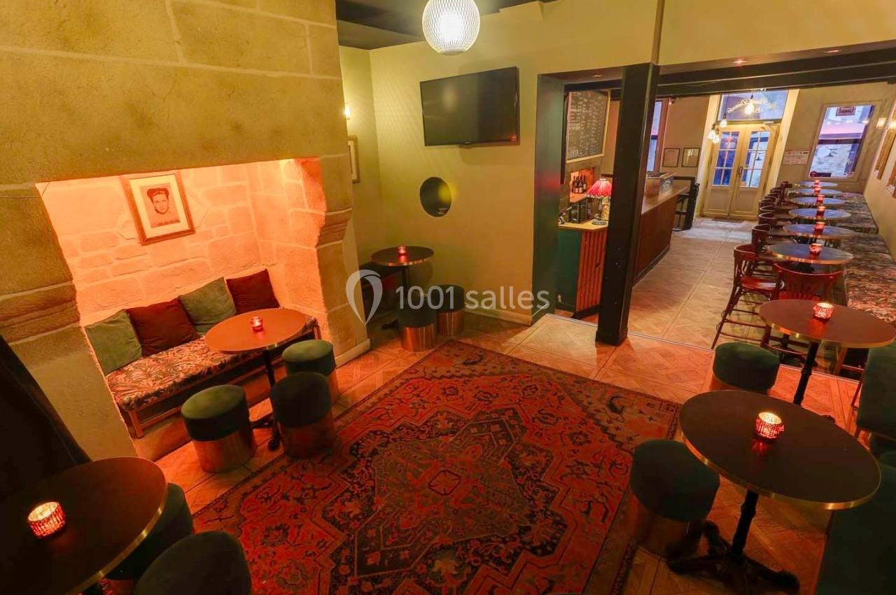 Salle de bar chaleureuse avec tables basses, banquettes, tapis rouge et lumière tamisée.