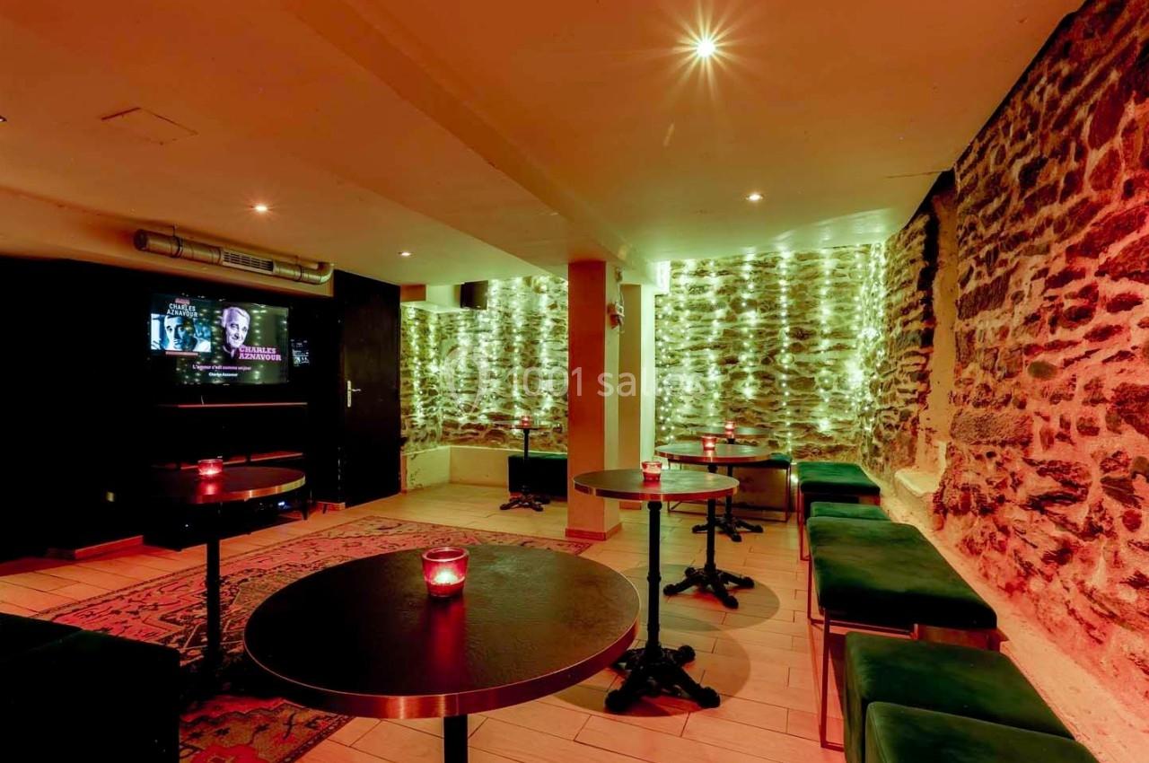 Salle avec murs en pierre, tables rondes noires, banquettes vertes et éclairage tamisé avec guirlandes lumineuses.