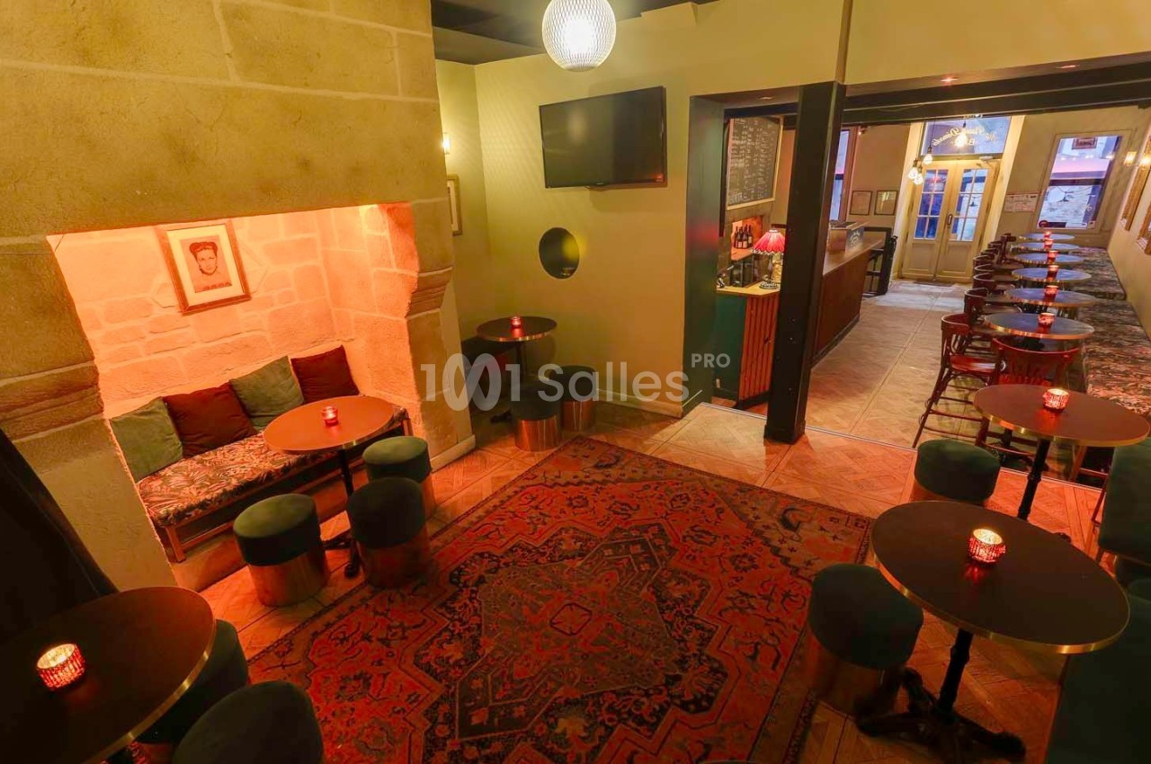 Salle de bar chaleureuse avec tables basses, banquettes, tapis rouge et lumière tamisée.