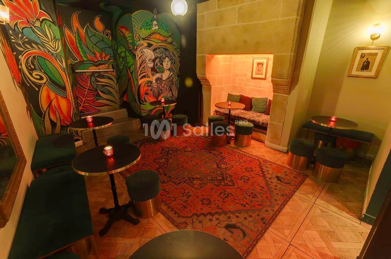 Salle cosy avec tables basses, tabourets verts, tapis rouge, fresques colorées et alcôve en pierre éclairée.