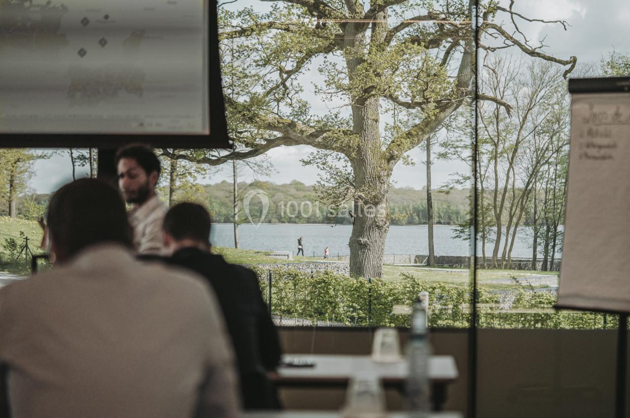 Photo Natura Parc #10 Vue depuis une salle de réunion avec des participants, donnant sur un lac entouré d'arbres et un chemin piéton.