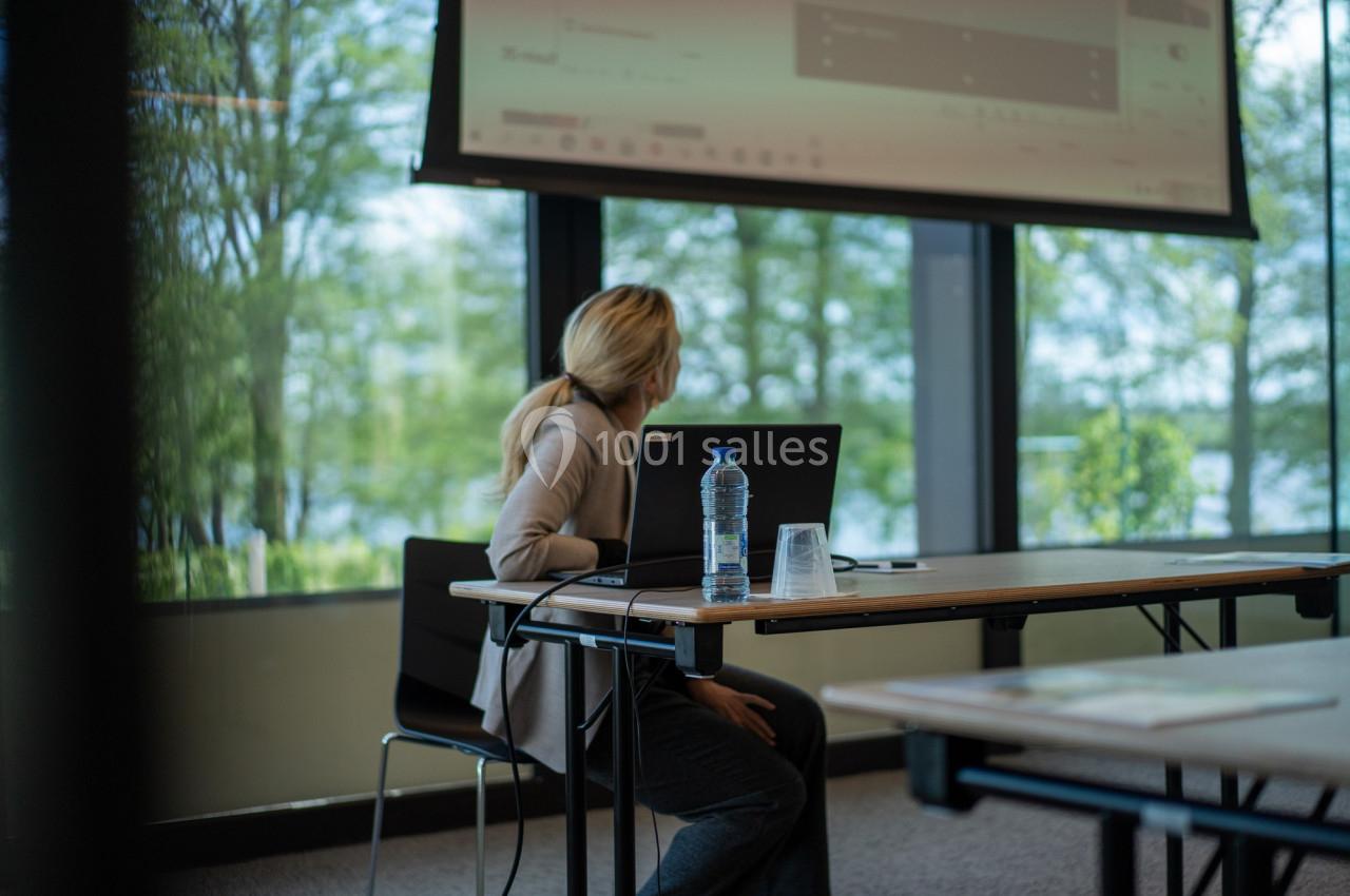 Photo Natura Parc #12 Une femme assise à une table regarde un écran de projection dans une salle lumineuse avec vue sur l'extérieur.