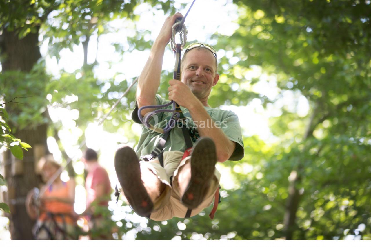 Photo Natura Parc #6 Un homme souriant descend en tyrolienne dans une forêt verdoyante par une journée ensoleillée.