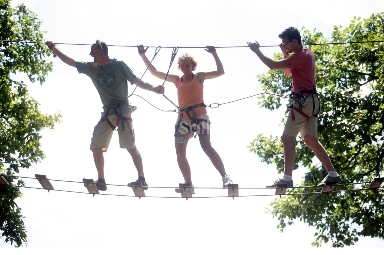 Trois personnes équipées de harnais traversent un pont suspendu en cordes dans un parcours d'accrobranche.