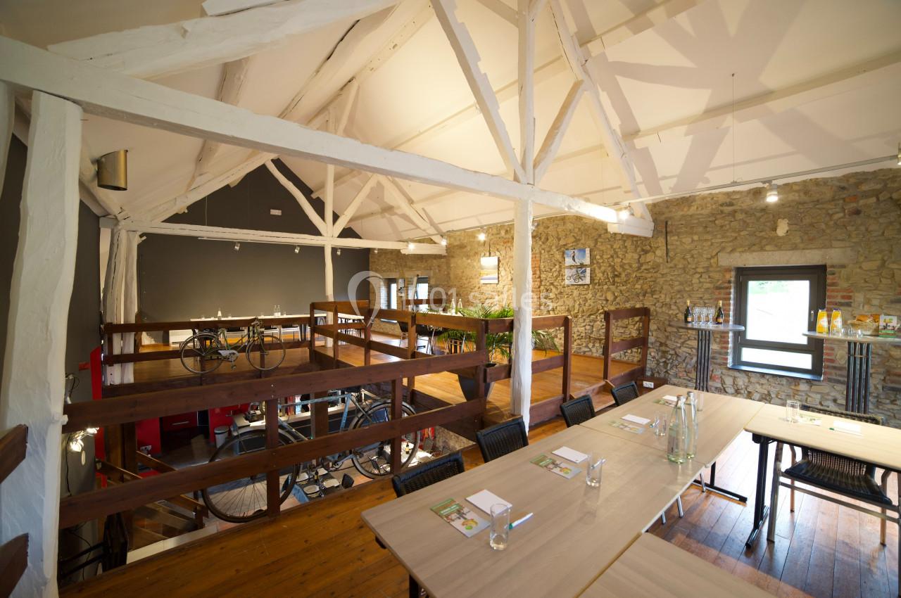 Photo Natura Parc #21 Salle lumineuse avec poutres apparentes, tables en bois, murs en pierre et mezzanine aménagée avec des vélos exposés.