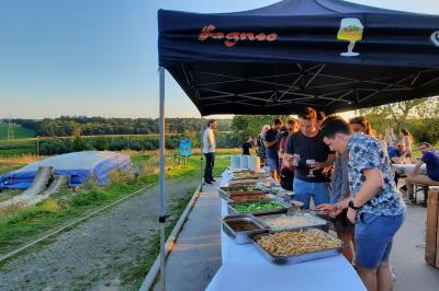 Miniature Natura Parc #25 Groupe de personnes rassemblées devant une maison en pierre avec un chapiteau, sur une pelouse par temps ensoleillé.