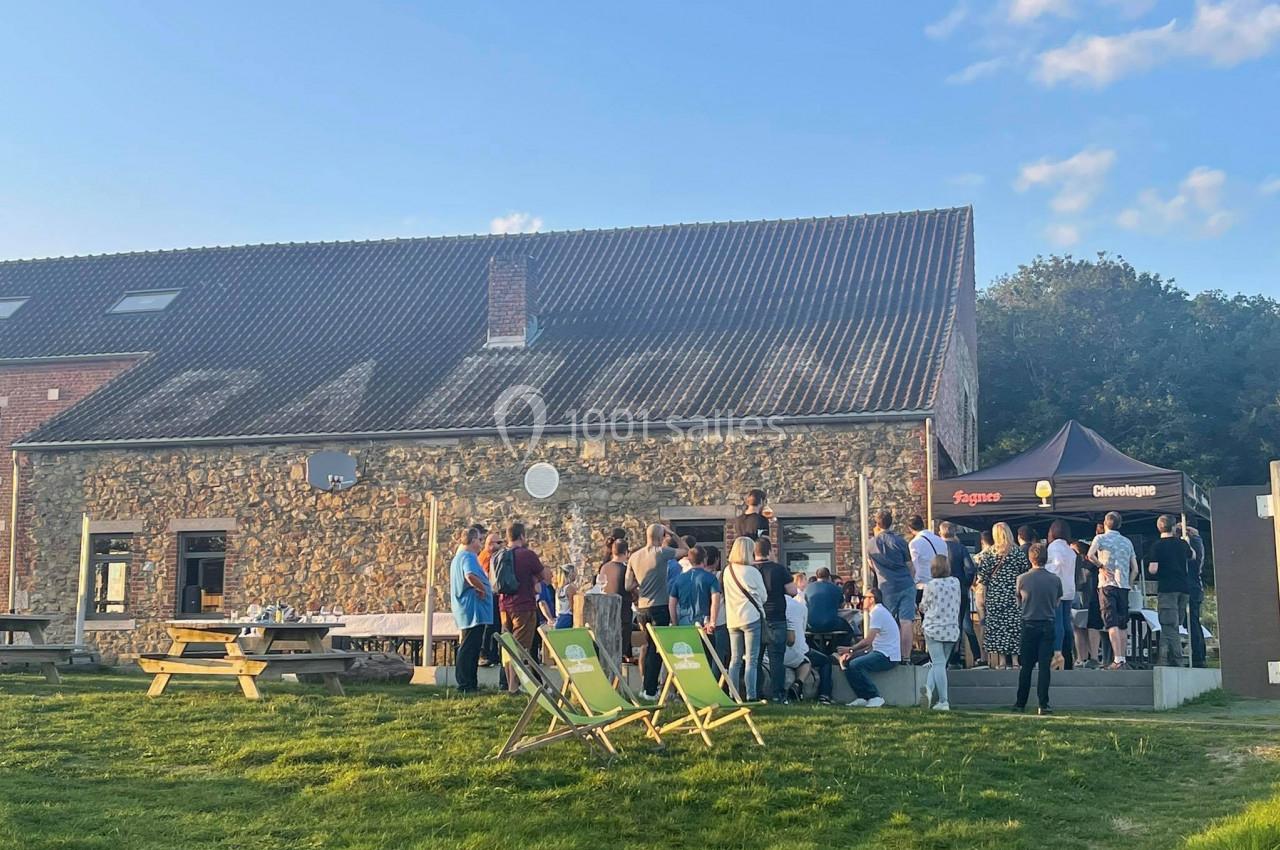 Photo Natura Parc #26 Groupe de personnes rassemblées devant une maison en pierre avec un chapiteau, sur une pelouse par temps ensoleillé.