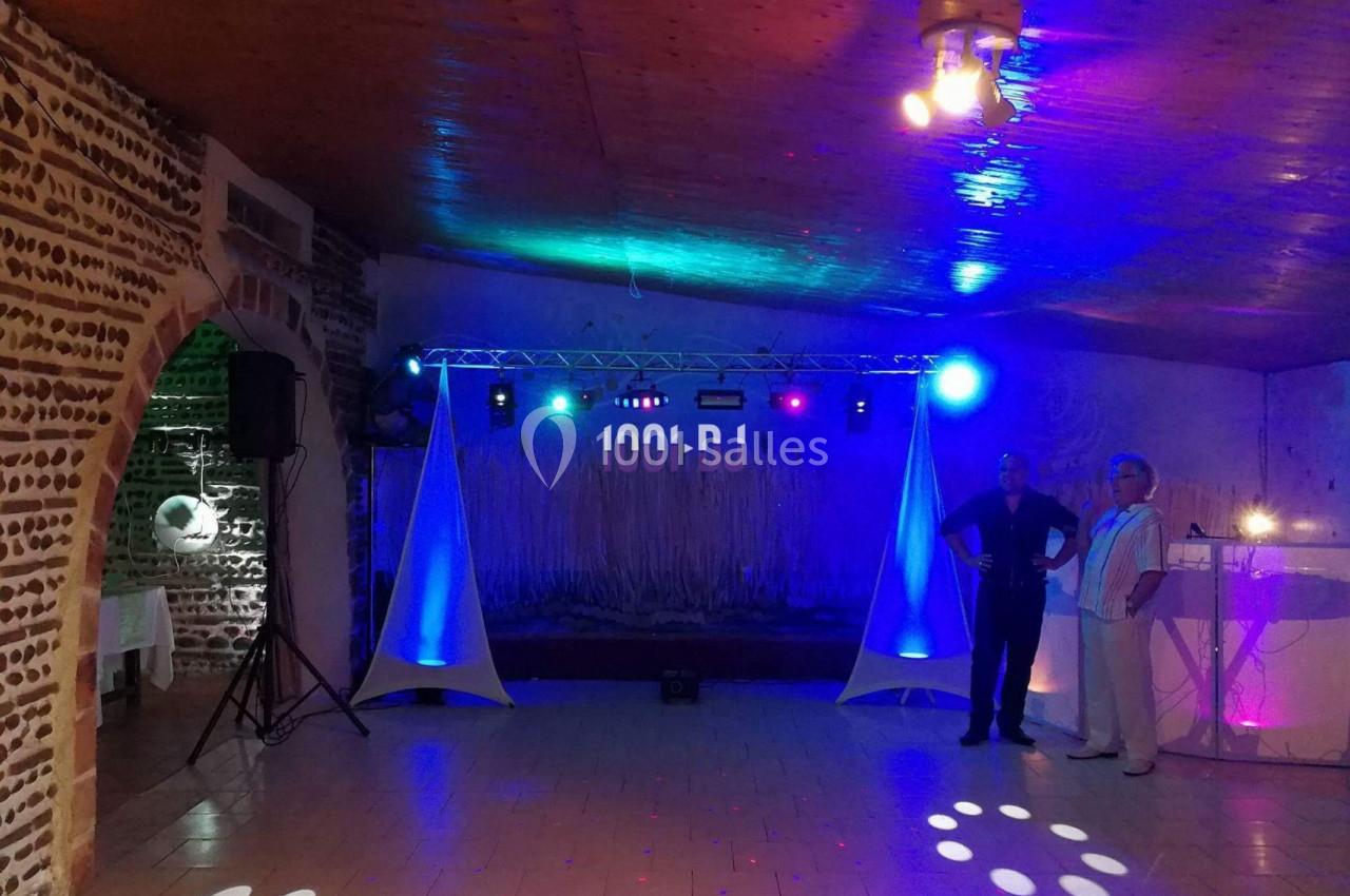 Salle de réception avec éclairage coloré, piste de danse vide et deux personnes debout près d'un bar lumineux.