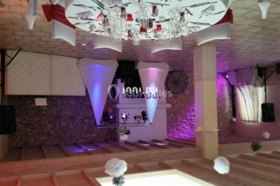 Salle de réception avec éclairage coloré, piste de danse vide et deux personnes debout près d'un bar lumineux.