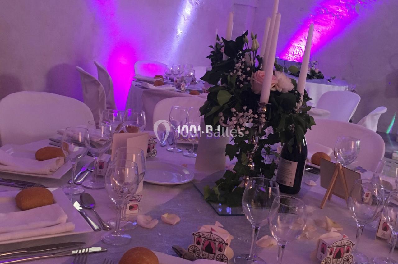 Table de réception décorée avec des bougies, des fleurs et des accessoires de mariage, dans une salle aux lumières violettes.