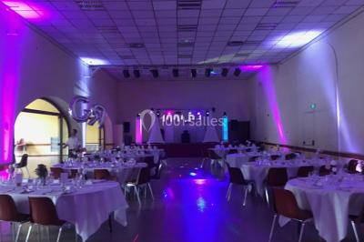 Salle de réception avec éclairage coloré, piste de danse vide et deux personnes debout près d'un bar lumineux.