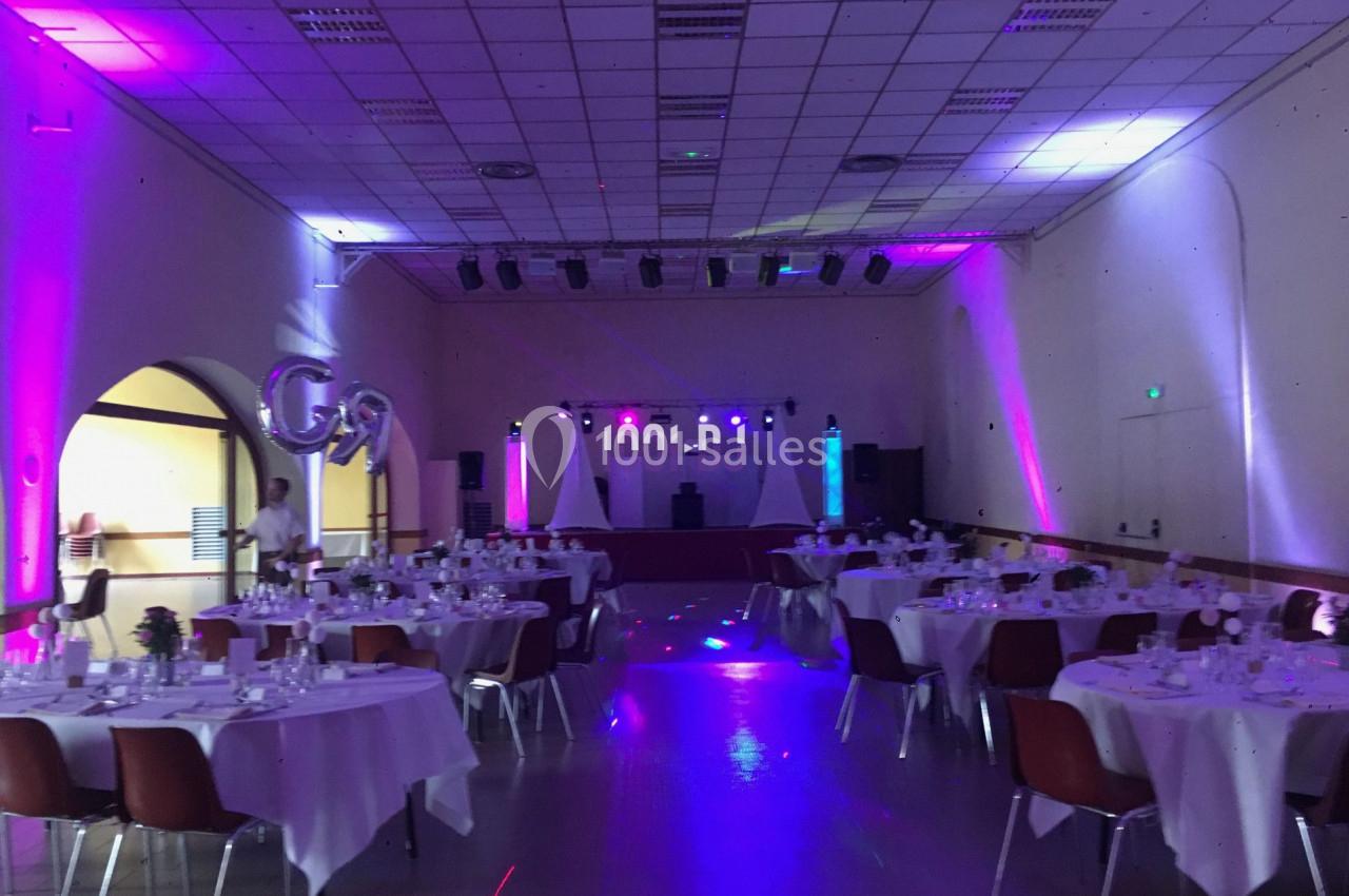 Salle de réception décorée avec des tables rondes dressées et une scène éclairée de lumières colorées.