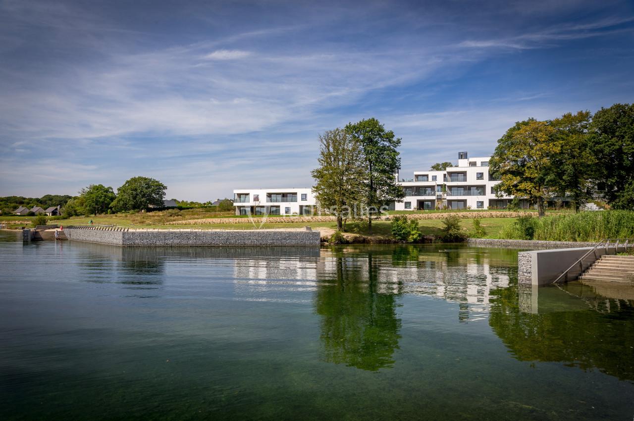 Vue d'un lac bordé de verdure avec des bâtiments modernes blancs en arrière-plan sous un ciel dégagé.