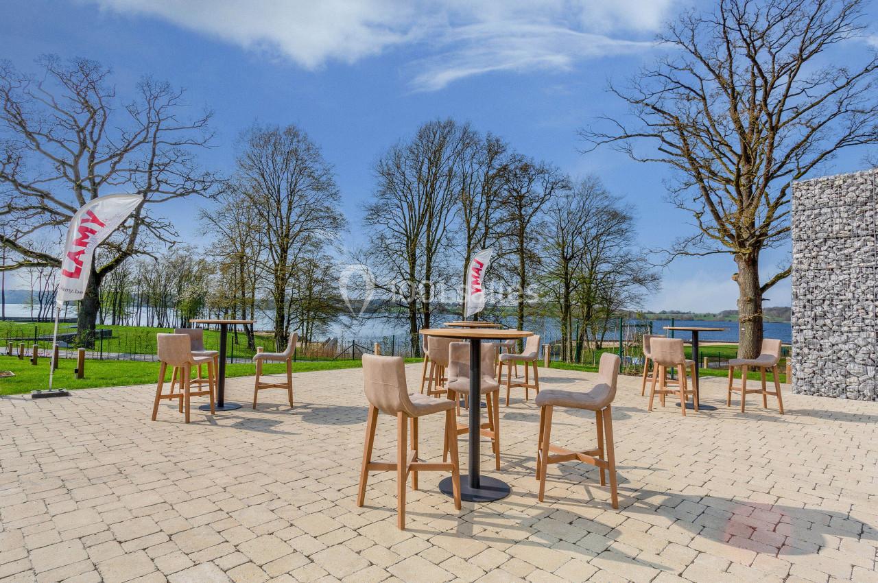 Terrasse extérieure avec tables hautes et chaises, entourée d'arbres et offrant une vue sur un plan d'eau.