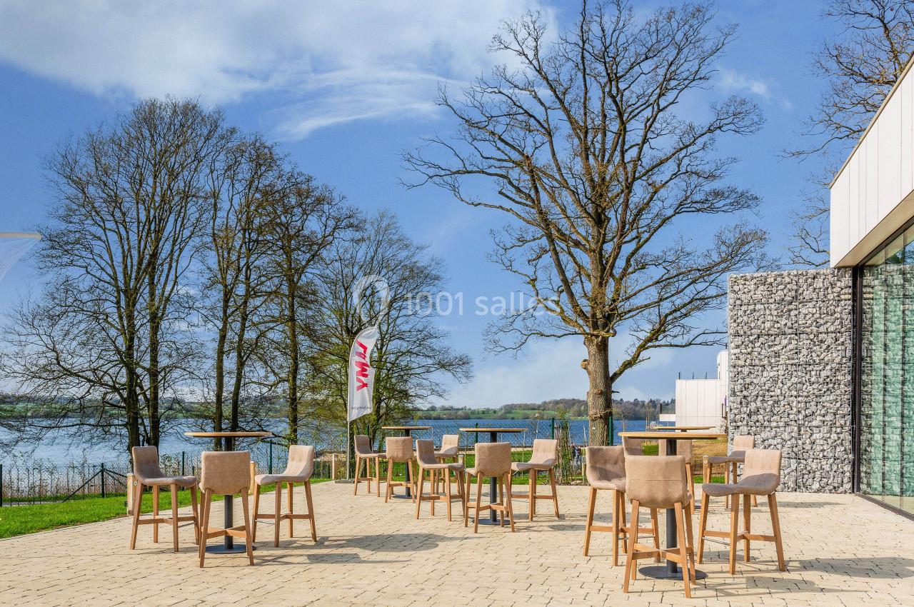 Terrasse extérieure avec tables et chaises, vue sur un lac entouré d'arbres sous un ciel partiellement nuageux.