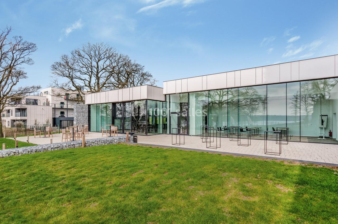 Bâtiment moderne avec façades vitrées donnant sur une terrasse et un espace vert, entouré d'arbres.