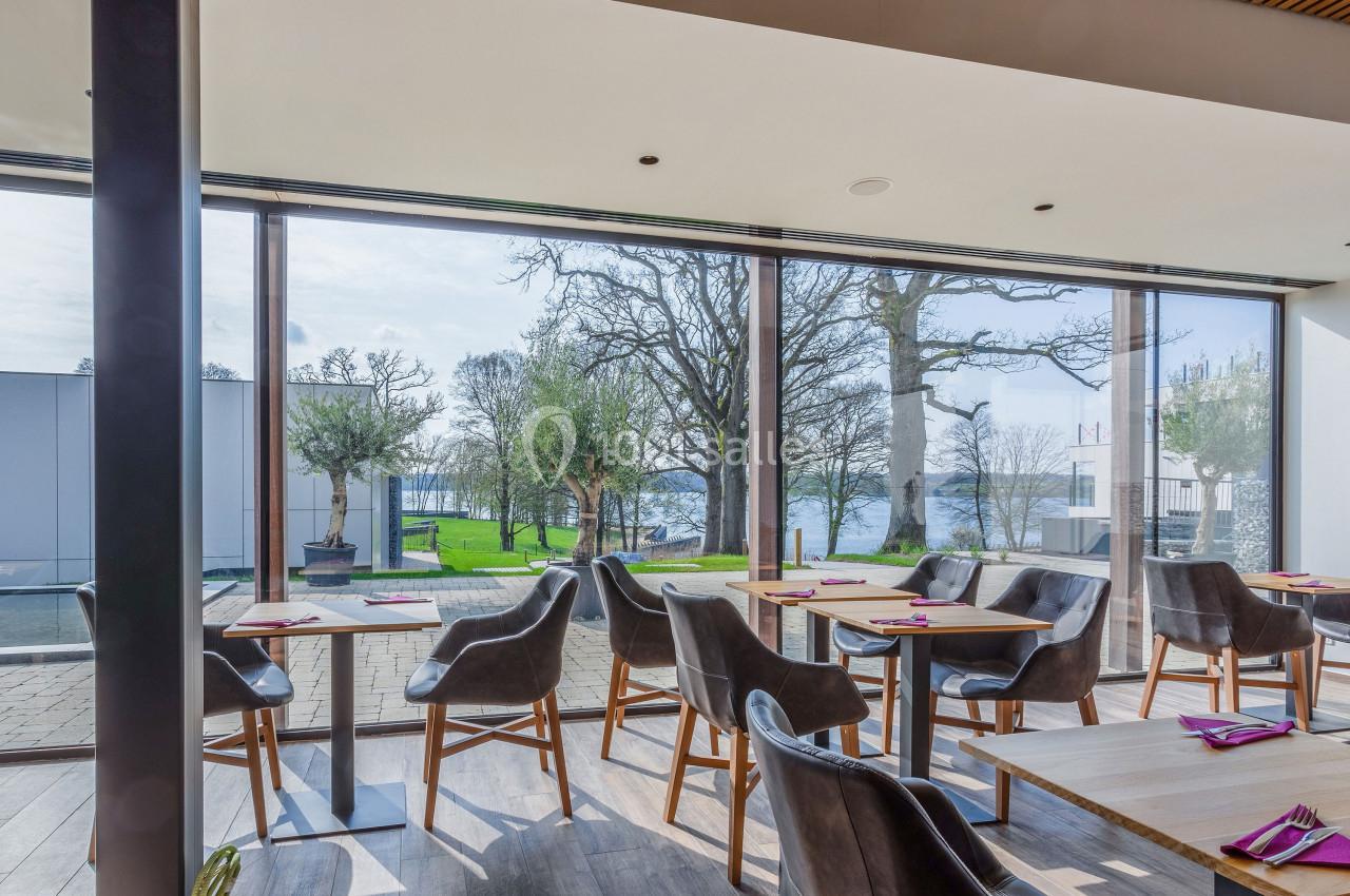 Salle de restaurant lumineuse avec grandes baies vitrées donnant sur un jardin et une vue sur un plan d'eau.