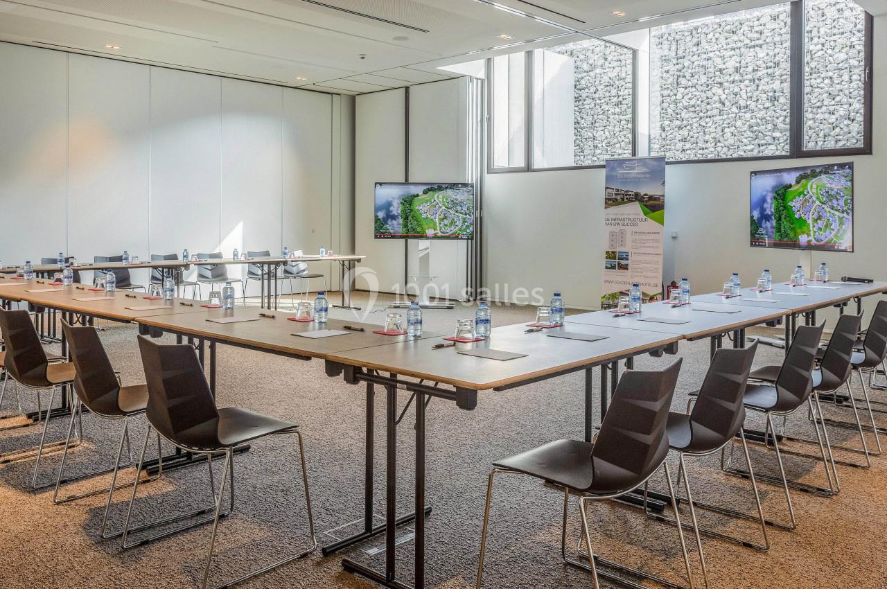Salle de réunion lumineuse avec tables disposées en U, chaises, écrans et documents sur les tables.