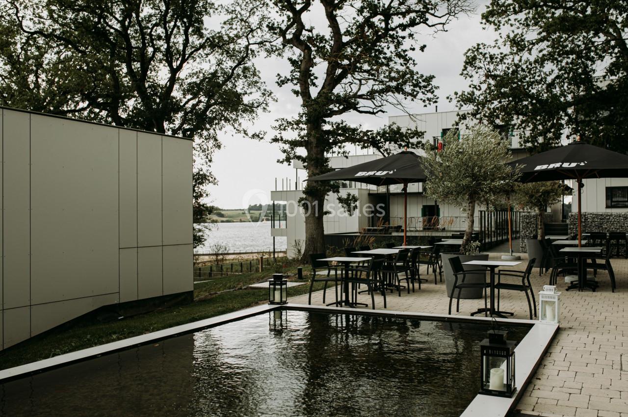 Terrasse extérieure avec tables, parasols et bassin d'eau, entourée d'arbres et donnant sur un paysage naturel.