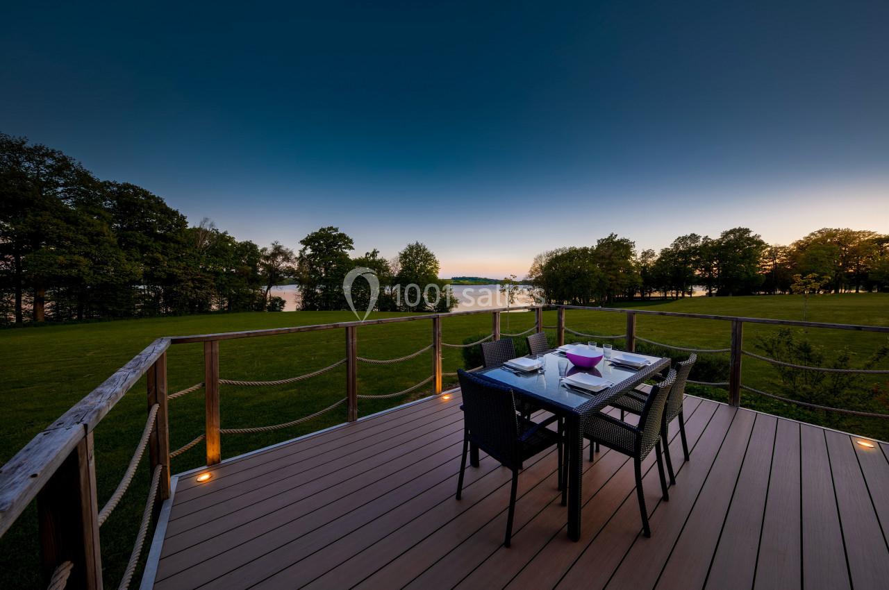 Terrasse en bois avec table et chaises, vue sur un jardin verdoyant et un lac au coucher du soleil.