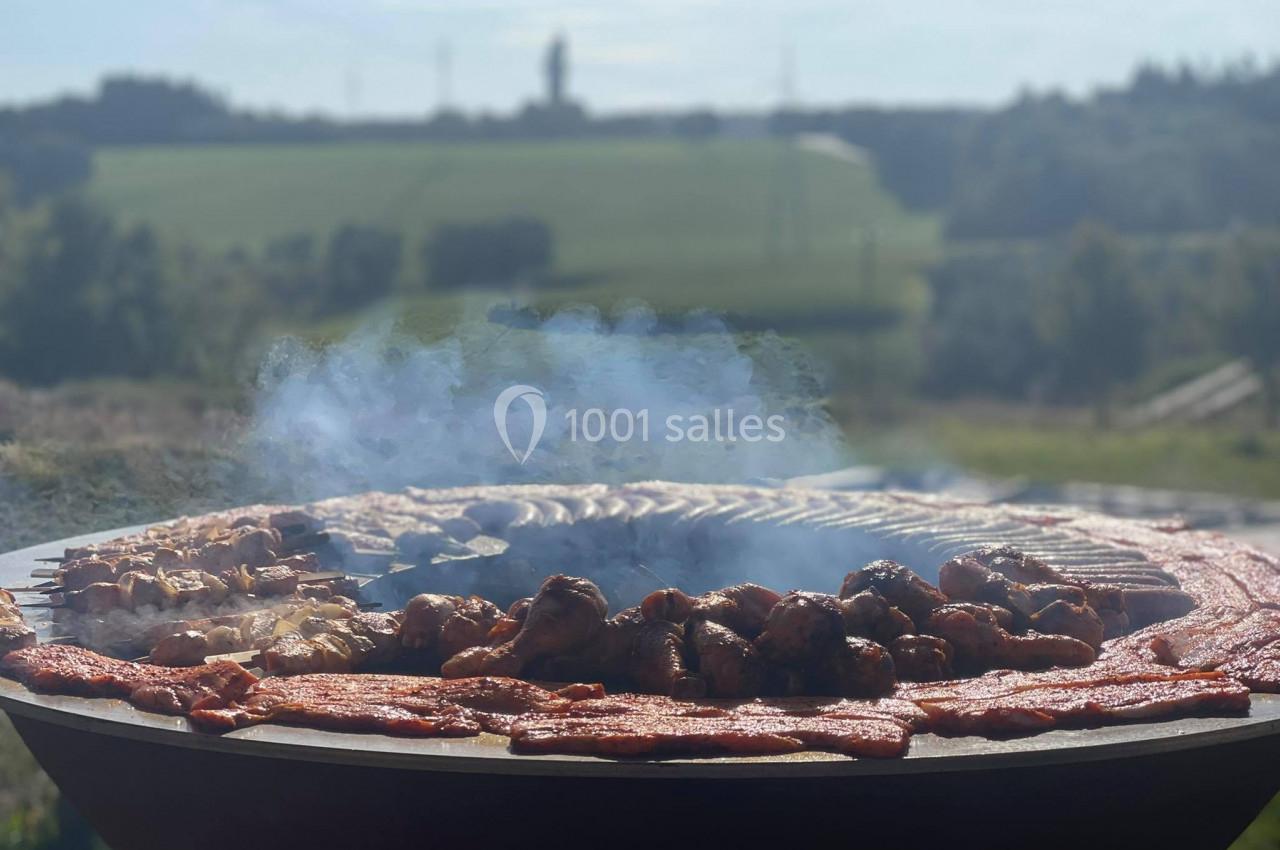 Viande variée grillant sur un barbecue circulaire en plein air, avec un paysage champêtre en arrière-plan.