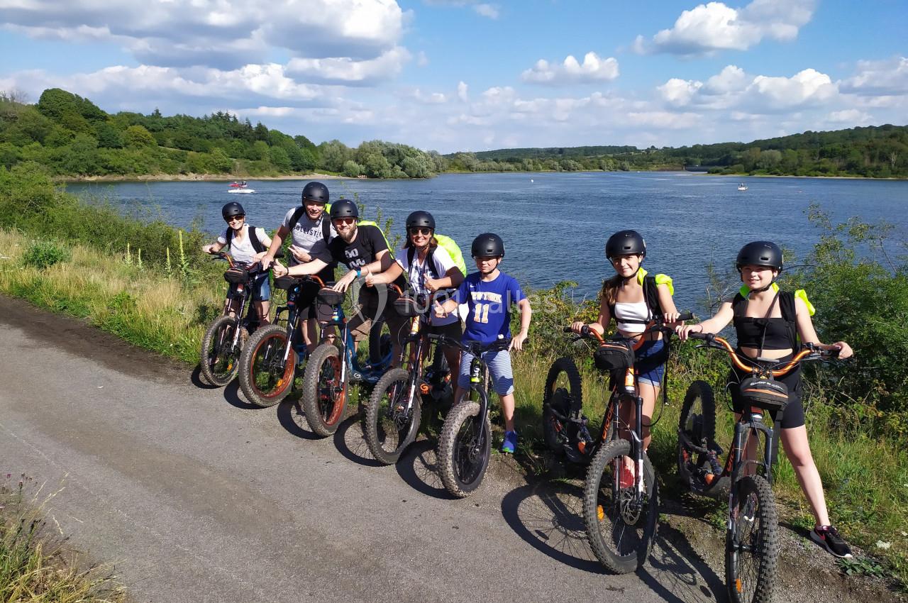 Un groupe de personnes portant des casques posent avec des trottinettes électriques près d'un lac sous un ciel dégagé.