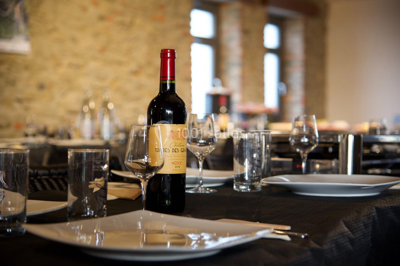 Bouteille de vin rouge posée sur une table dressée avec assiettes, verres et couverts, dans une salle lumineuse en pierre.