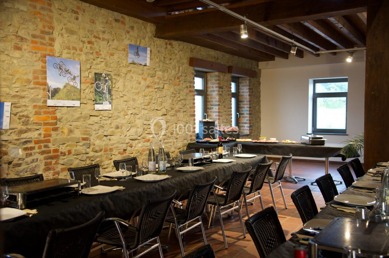 Salle à manger avec longues tables dressées, murs en pierre apparente, fenêtres et buffet en arrière-plan.