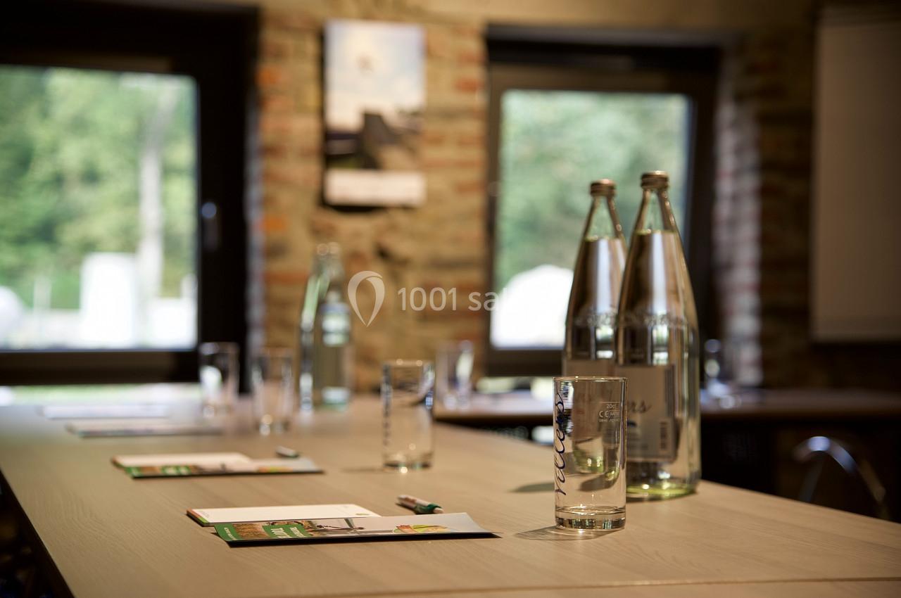 Salle de réunion avec une table en bois, des bouteilles d'eau, des verres et des documents devant des fenêtres ouvertes.