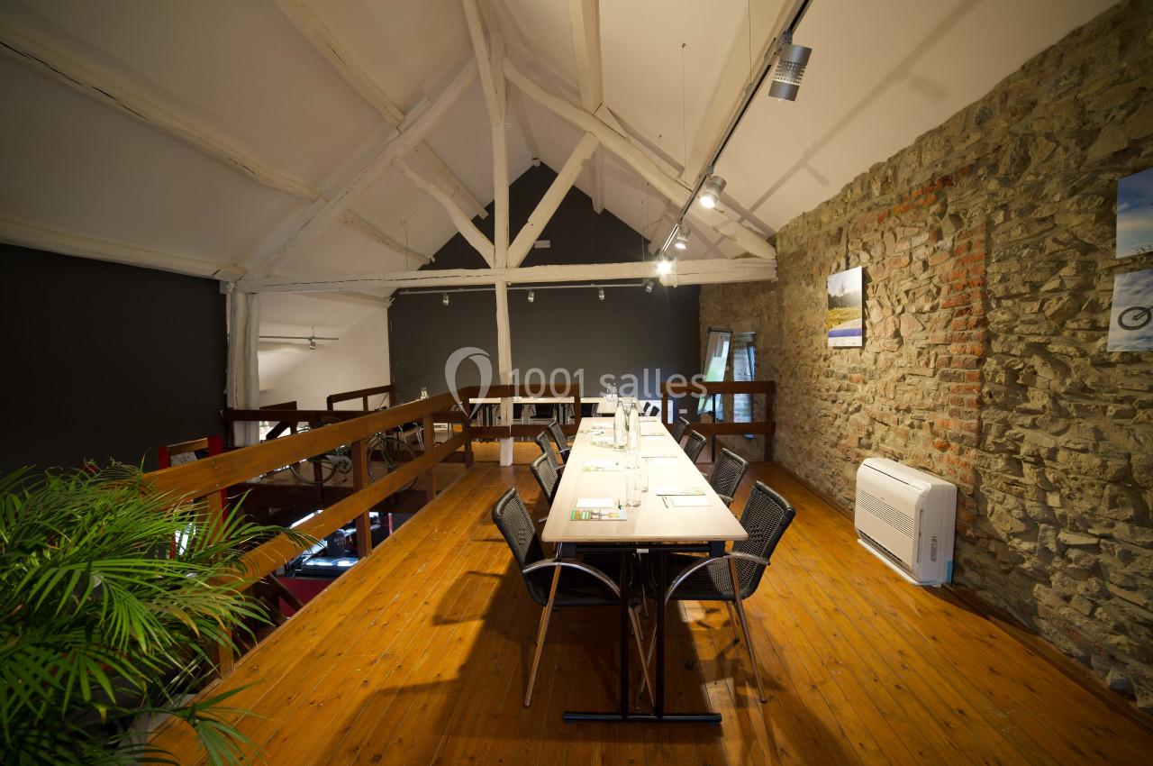 Salle de réunion lumineuse avec une grande table en bois, des chaises noires, murs en pierre et plafond avec poutres…
