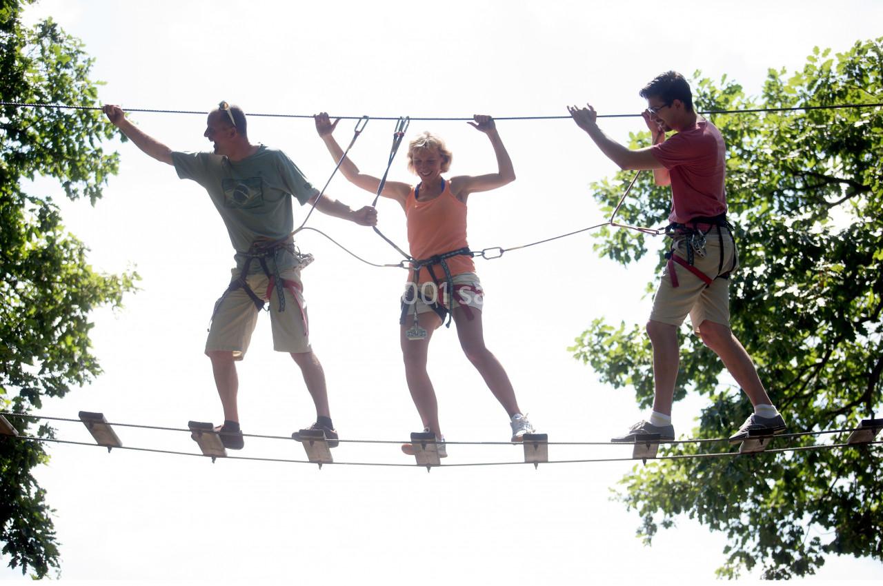 Trois personnes équipées de harnais traversent un pont suspendu en bois dans un parcours d'accrobranche en plein air.