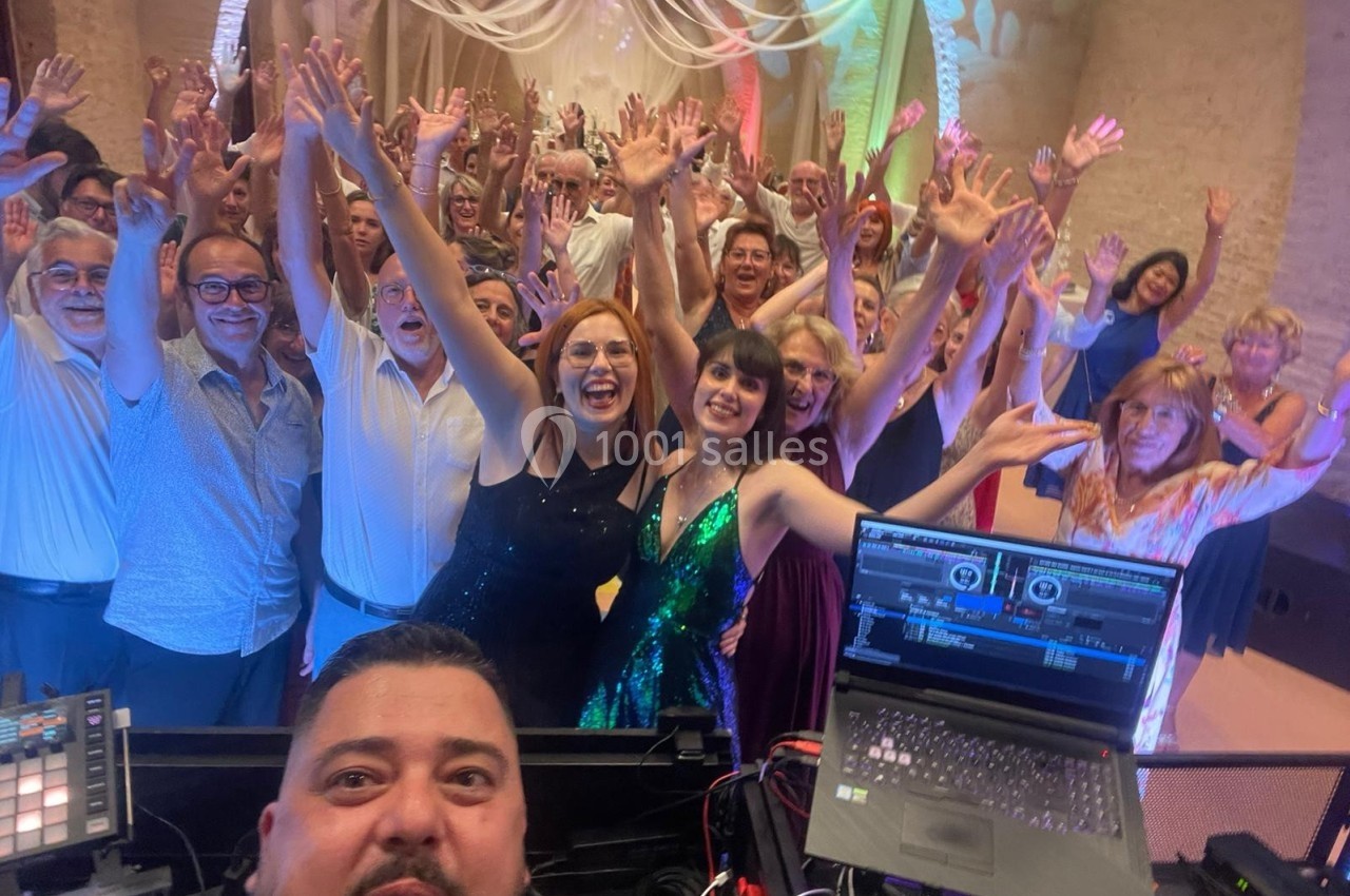 Un groupe de personnes souriantes levant les bras lors d'une fête, avec un DJ au premier plan et une ambiance festive.