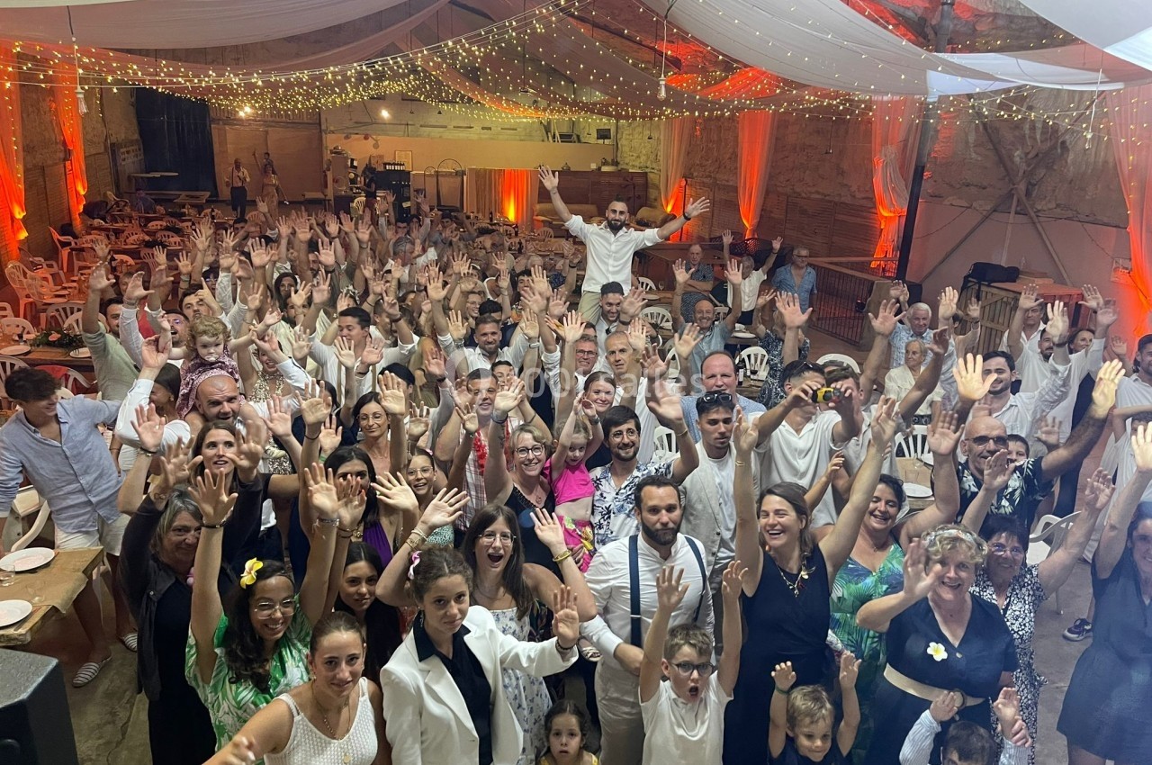 Groupe de personnes souriantes levant les bras dans une grande salle décorée de guirlandes lumineuses et de drapés blancs.