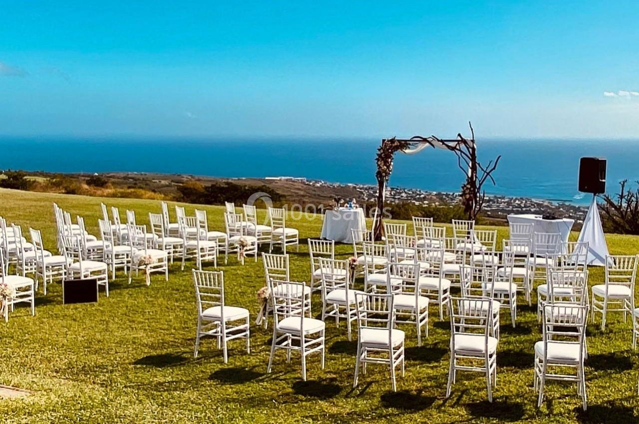 Chaises blanches disposées en extérieur sur une pelouse avec une arche décorée, vue sur la mer en arrière-plan.