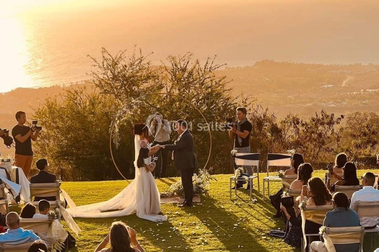 Un couple échange ses vœux de mariage en plein air au coucher du soleil, entouré d'invités assis.