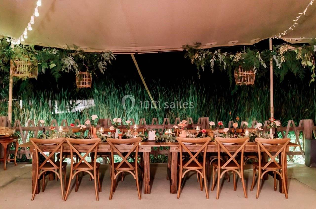 Table en bois décorée de fleurs et de bougies, sous une tente éclairée, avec des plantes en arrière-plan.