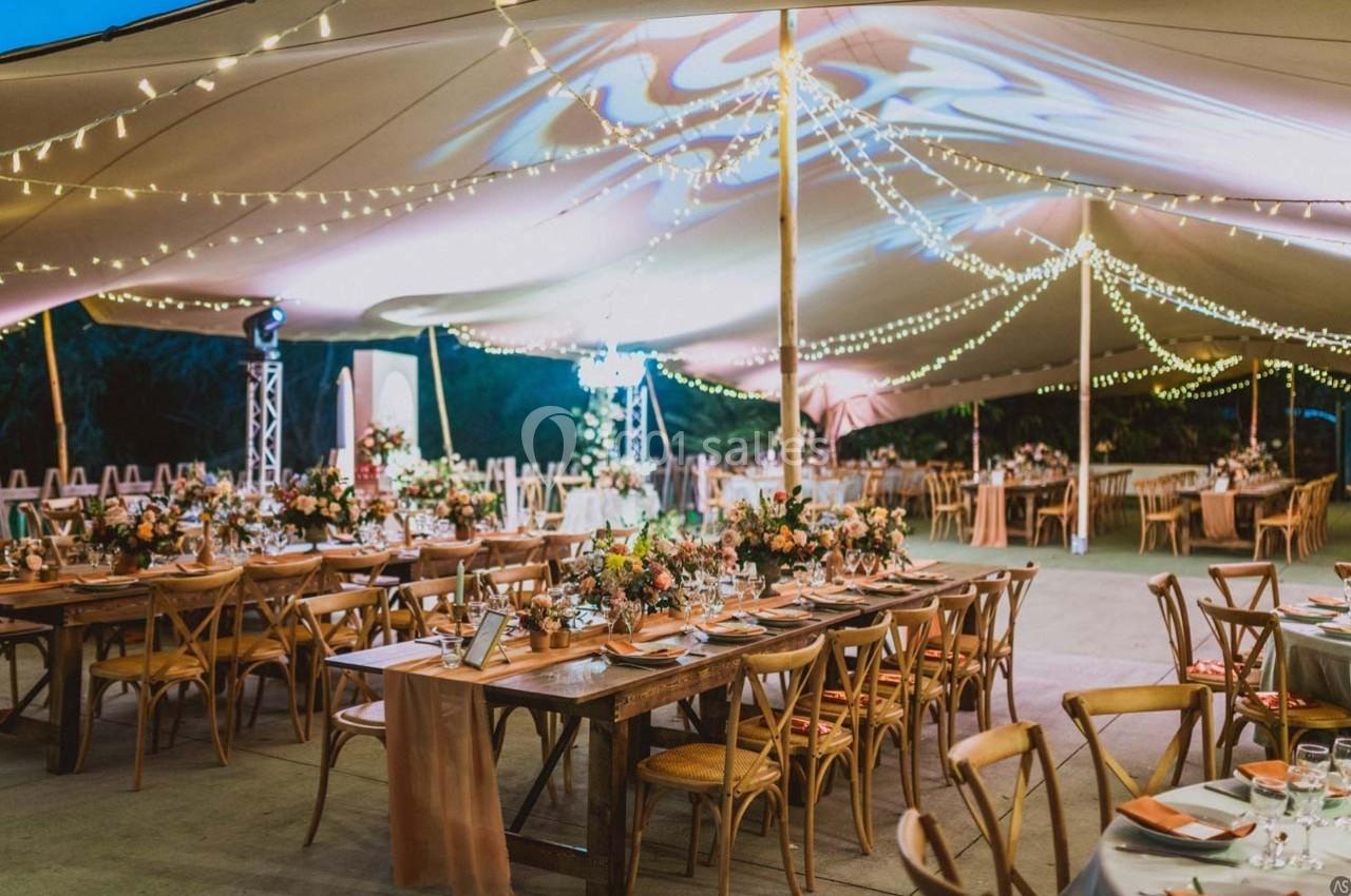 Salle de réception en plein air décorée avec des guirlandes lumineuses, tables en bois et arrangements floraux.