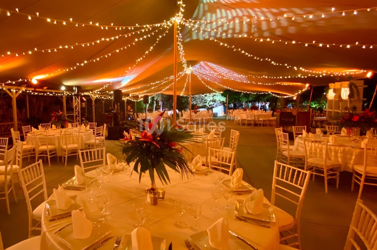 Salle de réception sous une tente illuminée avec des guirlandes, tables dressées et décorations florales au centre.
