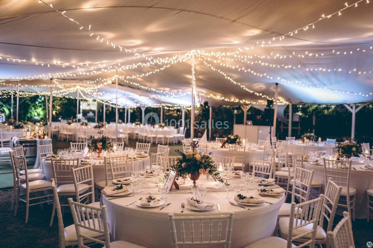 Salle de réception en plein air décorée de guirlandes lumineuses, avec des tables rondes dressées et des chaises blanches.