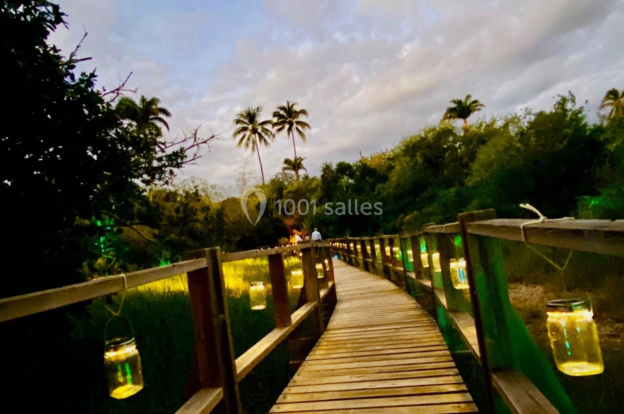 Passerelle en bois éclairée par des lanternes suspendues, traversant une végétation luxuriante au crépuscule.