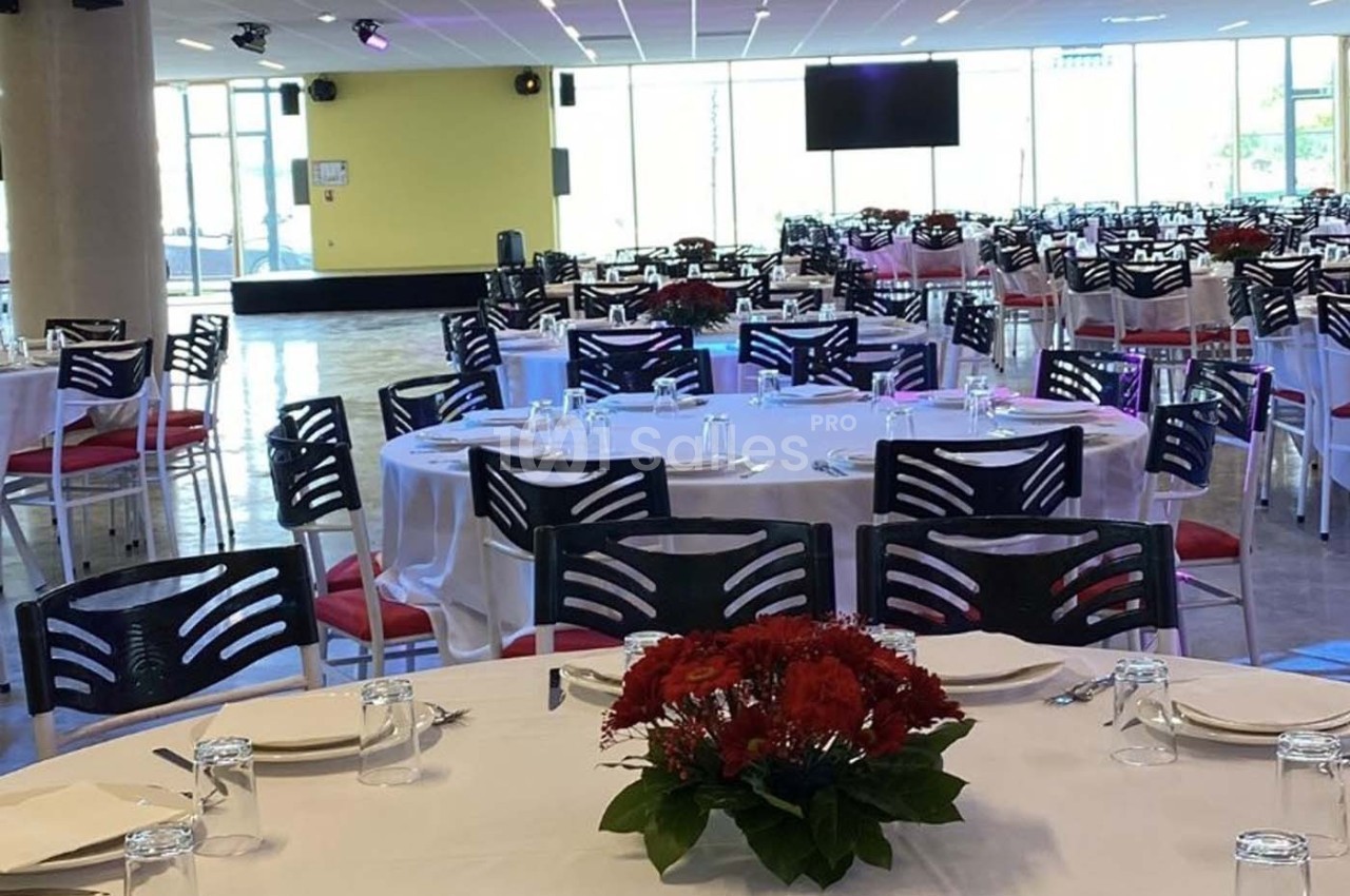 Salle de réception avec tables rondes dressées, nappes blanches, chaises noires et centres de table fleuris rouges.