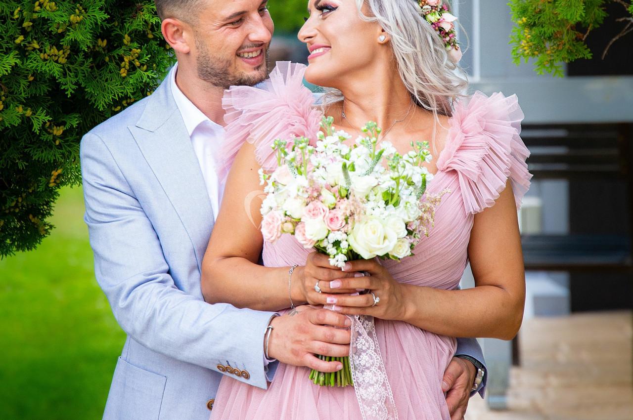 Un couple souriant en tenue élégante, la femme tenant un bouquet de fleurs, dans un jardin verdoyant.