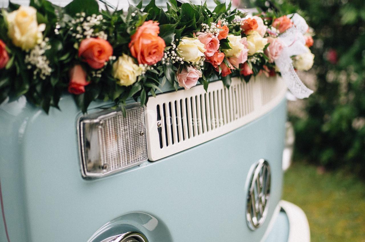 Van vintage bleu décoré d'une guirlande de fleurs colorées, garé près d'arbustes fleuris.